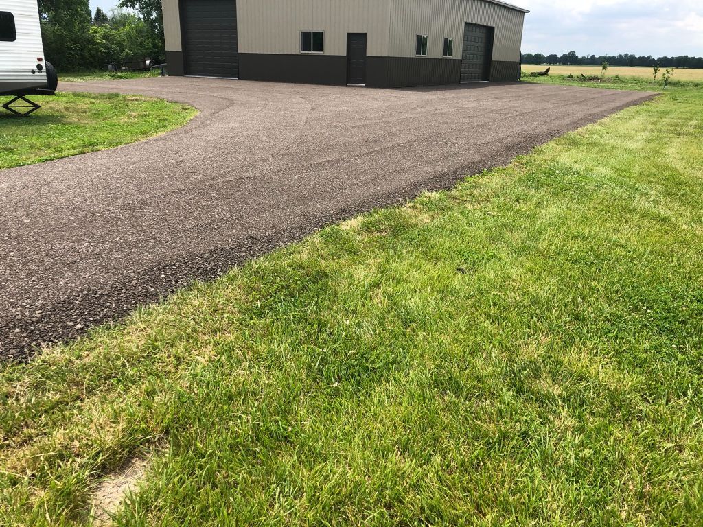 Gravel driveway beside a tan metal building, bordered by green grass under a clear sky