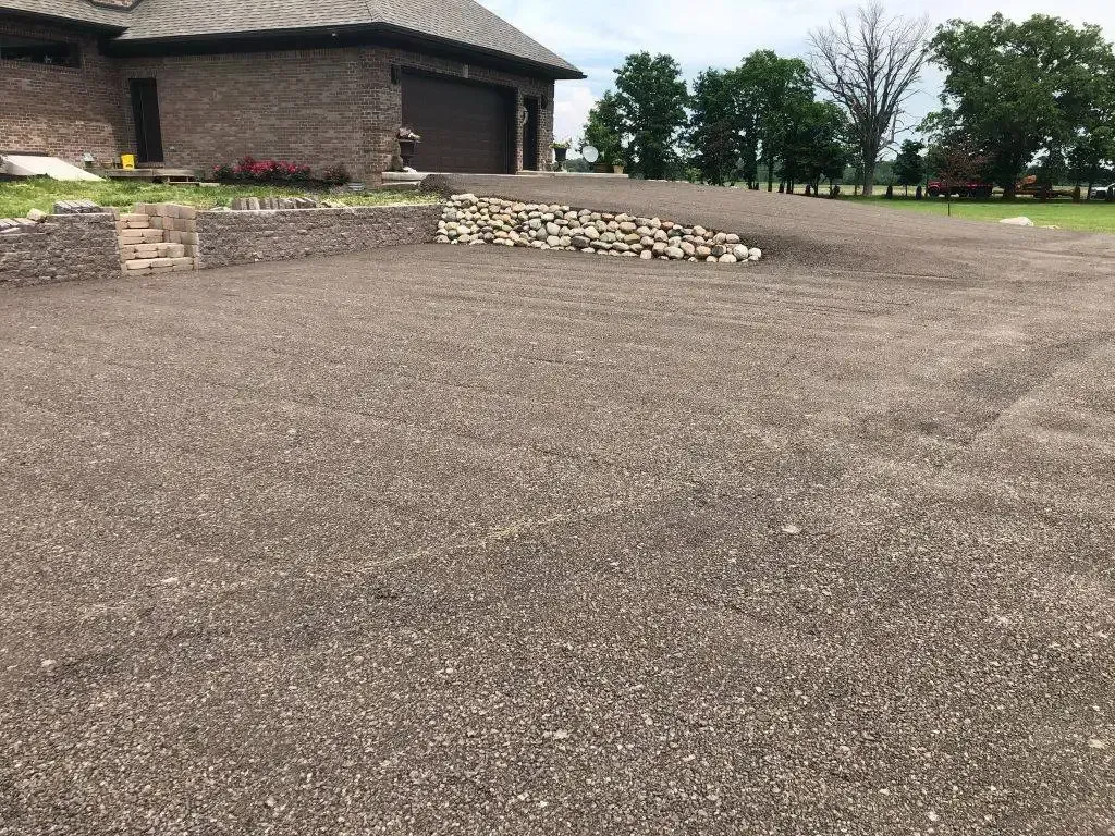 Gravel driveway beside a stone house with a low rock border and trees in the distance