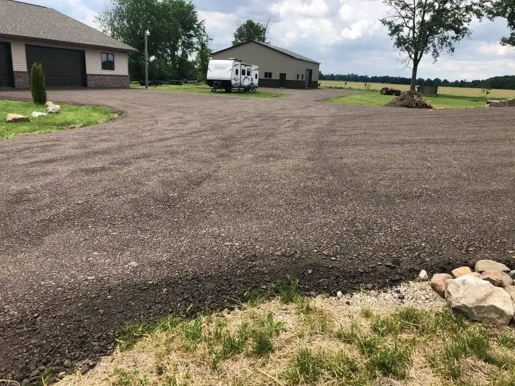 Gravel driveway leading to a house and garage in a rural yard, with grass and trees nearby