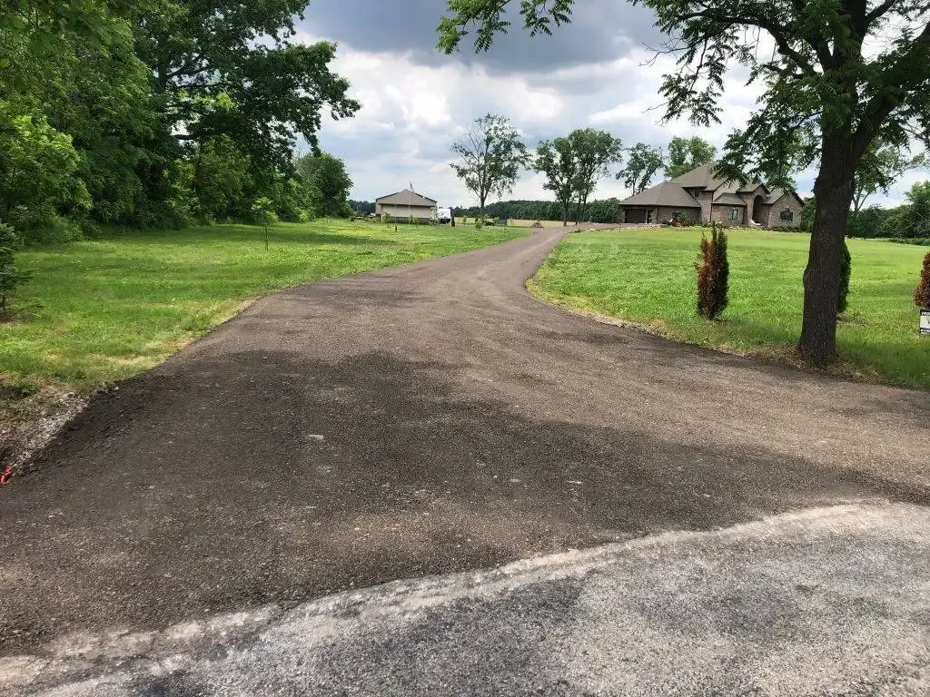 Gravel driveway curving through a green lawn toward houses under a cloudy sky.