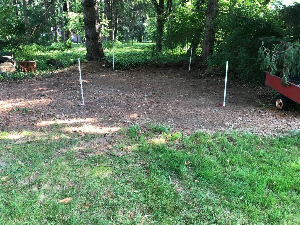 Backyard garden bed with bare soil, white stakes, and grass under trees