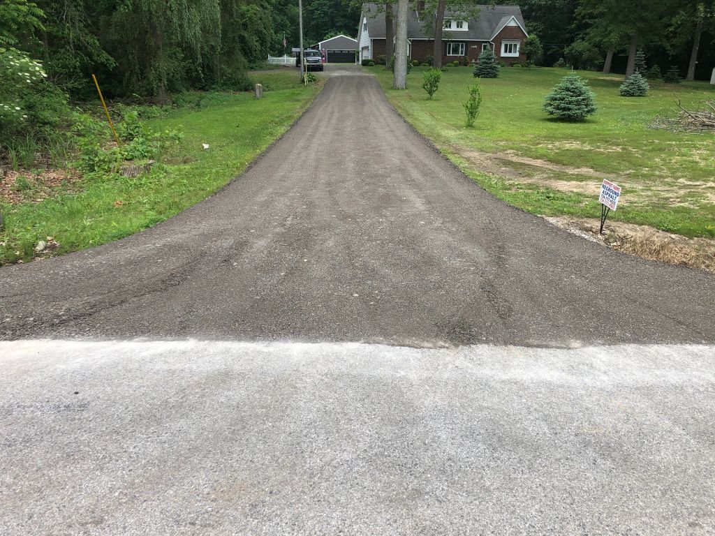 Gravel driveway leading to a house, bordered by green lawns and trees, with a mailbox on the right.