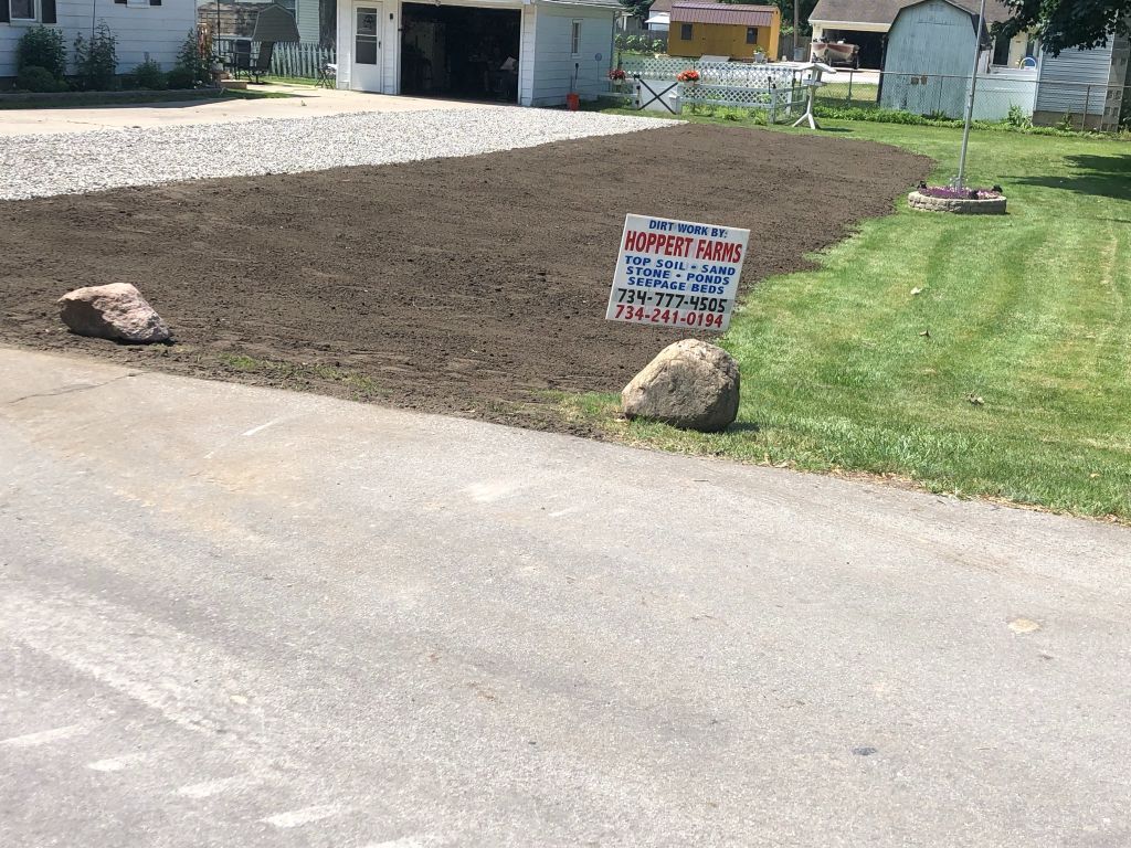 Freshly tilled garden bed beside a driveway, marked with a sign and bordered by rocks and lawn