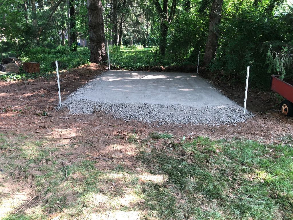 Concrete slab in a wooded area, surrounded by gravel and dirt with a white marker post.