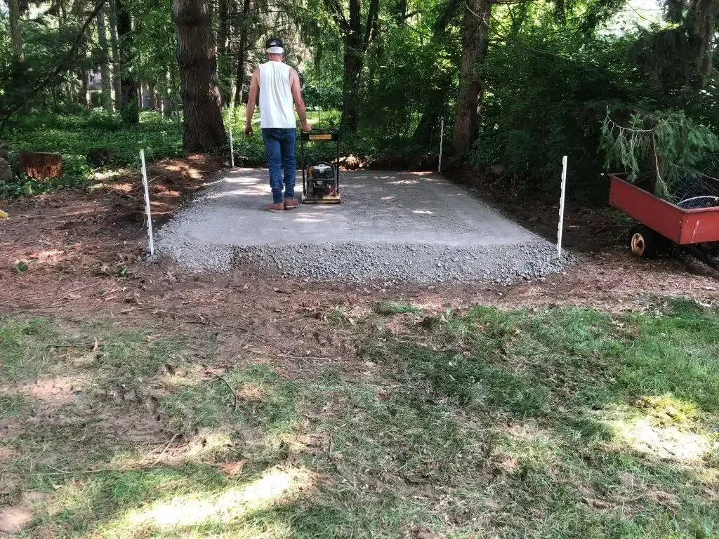 Person standing on a concrete slab in a wooded backyard with a small machine nearby.