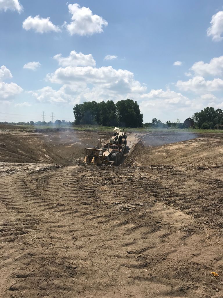 Excavator grading a dirt field under a blue sky with clouds, trees in the distance