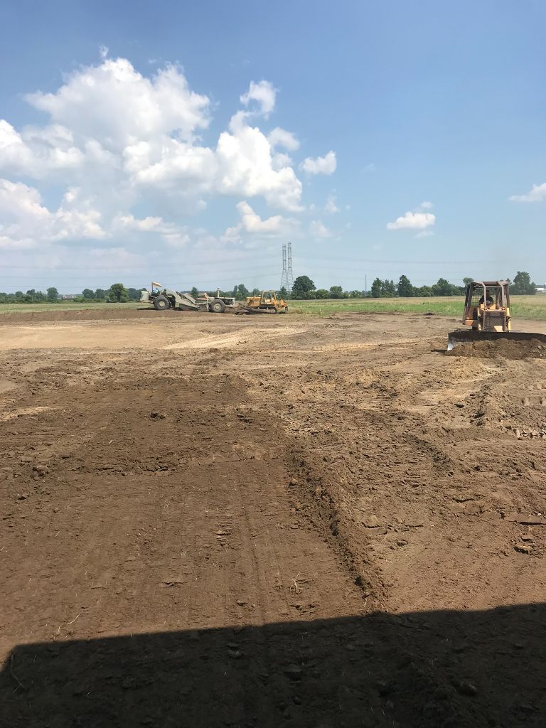 Wide dirt construction site under a blue sky, with a backhoe on the right and distant trees on the horizon.