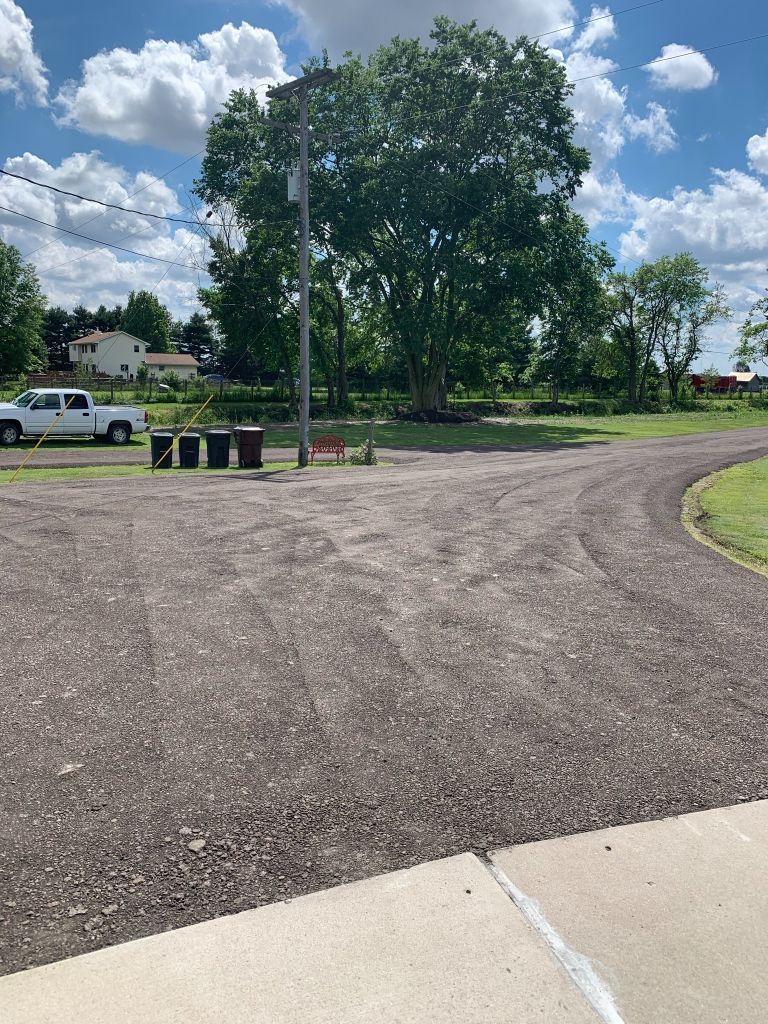 Gravel parking lot curving around a large tree under a bright blue sky with clouds