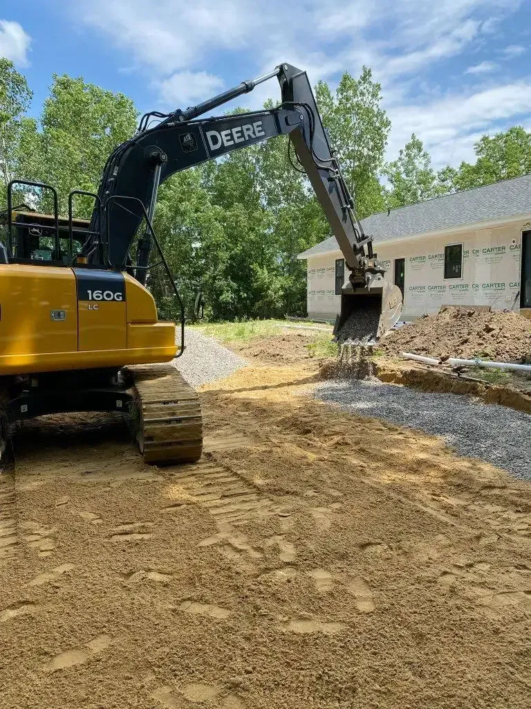 Yellow John Deere excavator demolishing a house in a dirt lot beside trees and a partly standing brick home