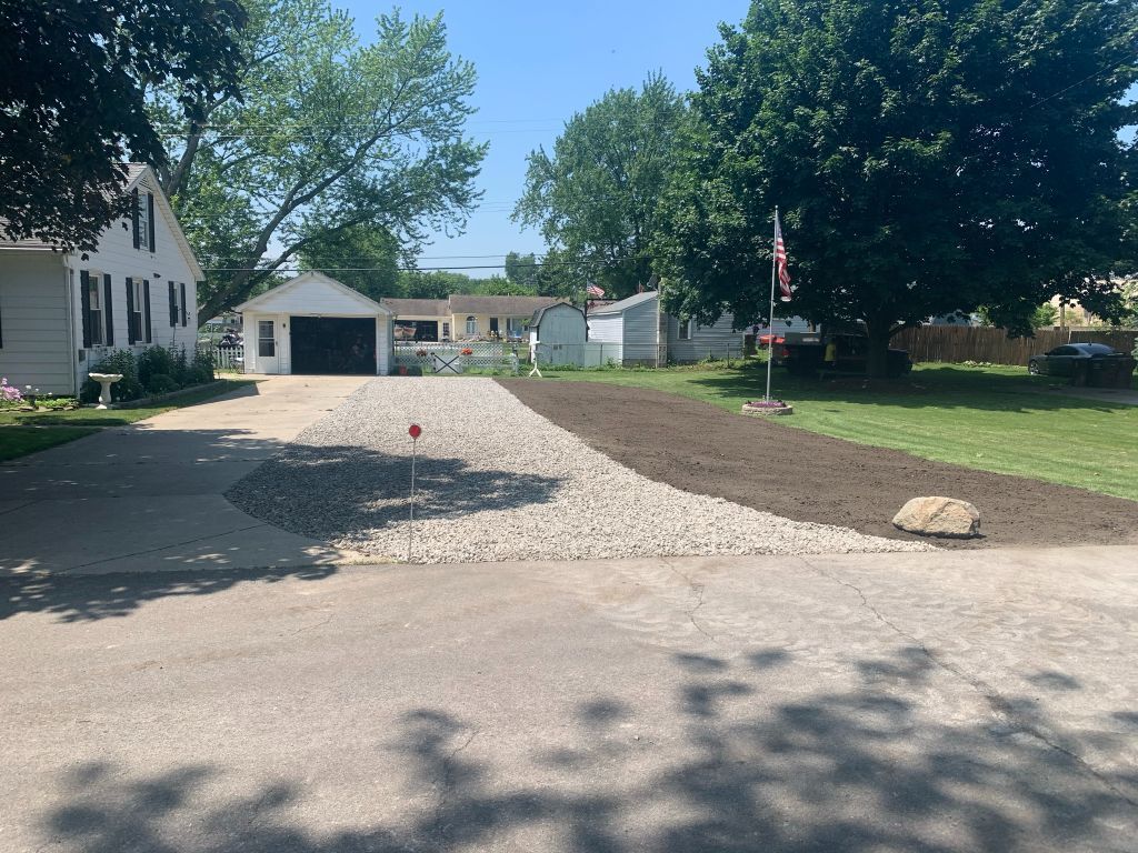 Gravel driveway leading to a house and garage, with a large tree and American flag on a sunny day