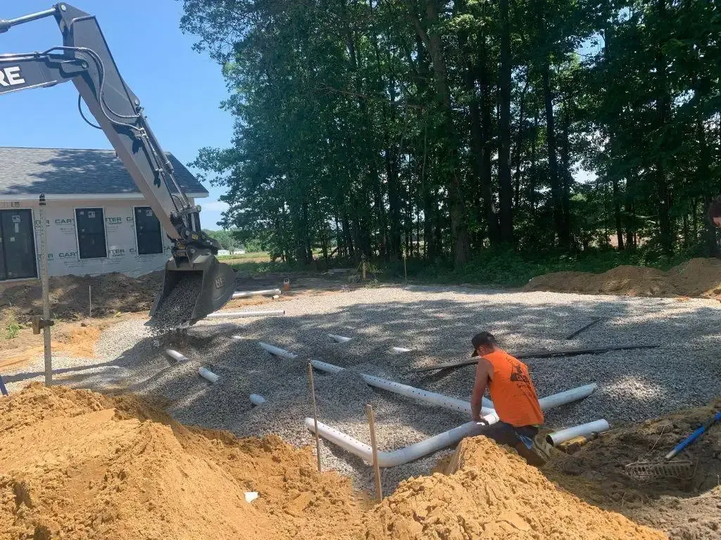 Excavator digging a trench beside a house, with a worker in an orange vest on a construction site.