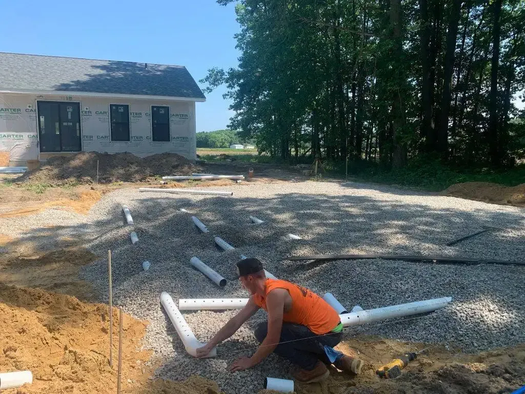 Worker laying white PVC pipes in a gravel trench beside a house under construction.