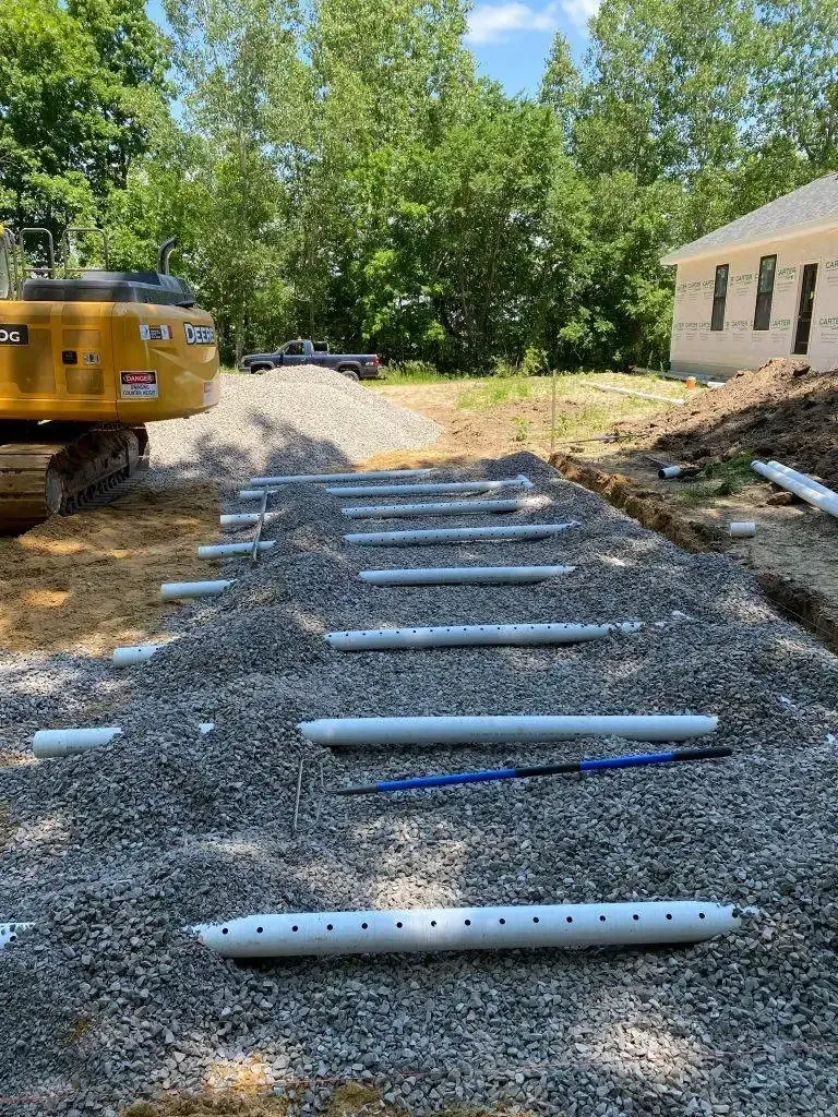 Gravel driveway under construction with metal pipes laid out, beside a yellow truck and a house.