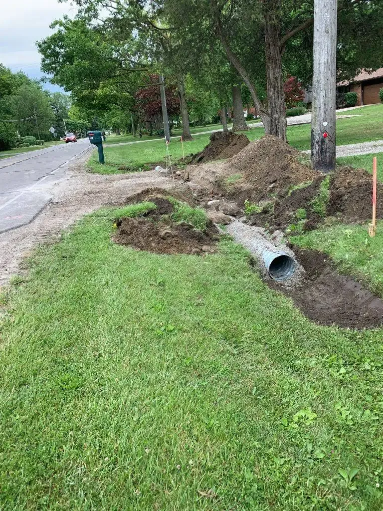 Roadside ditch collapse with exposed drainage pipe beside a grassy street and trees.