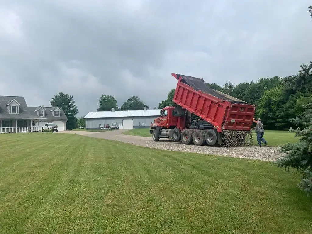 Red dump truck unloading gravel on a driveway beside a house and lawn under cloudy skies