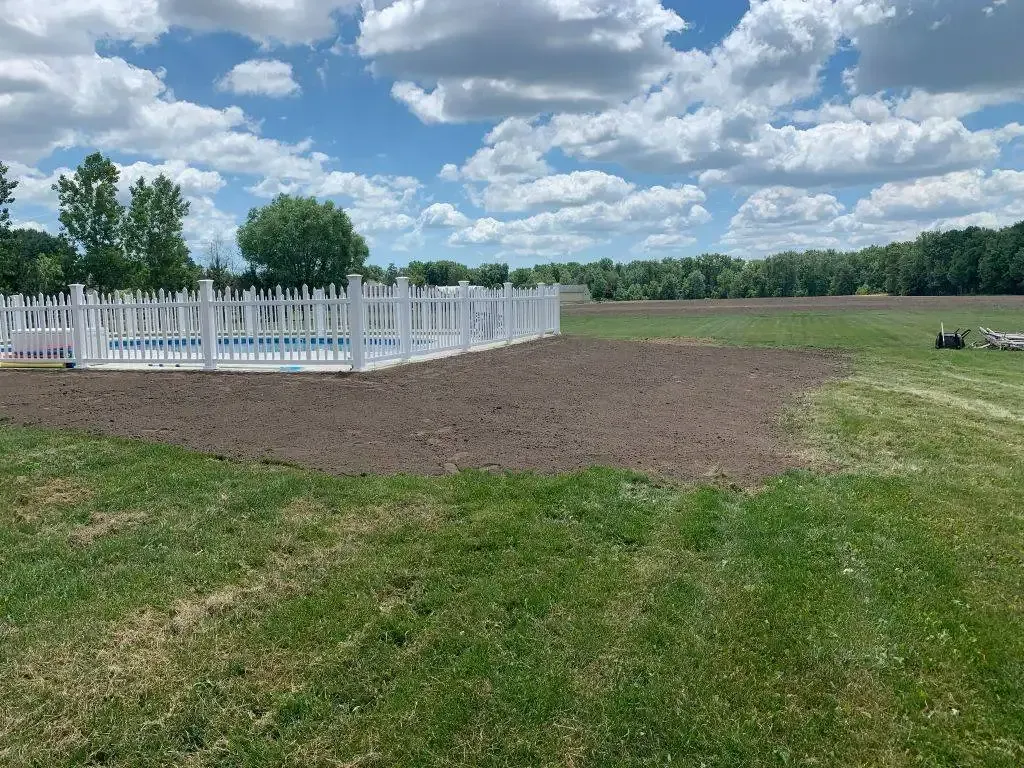 White picket fence around a small pool on a grassy field under a cloudy blue sky