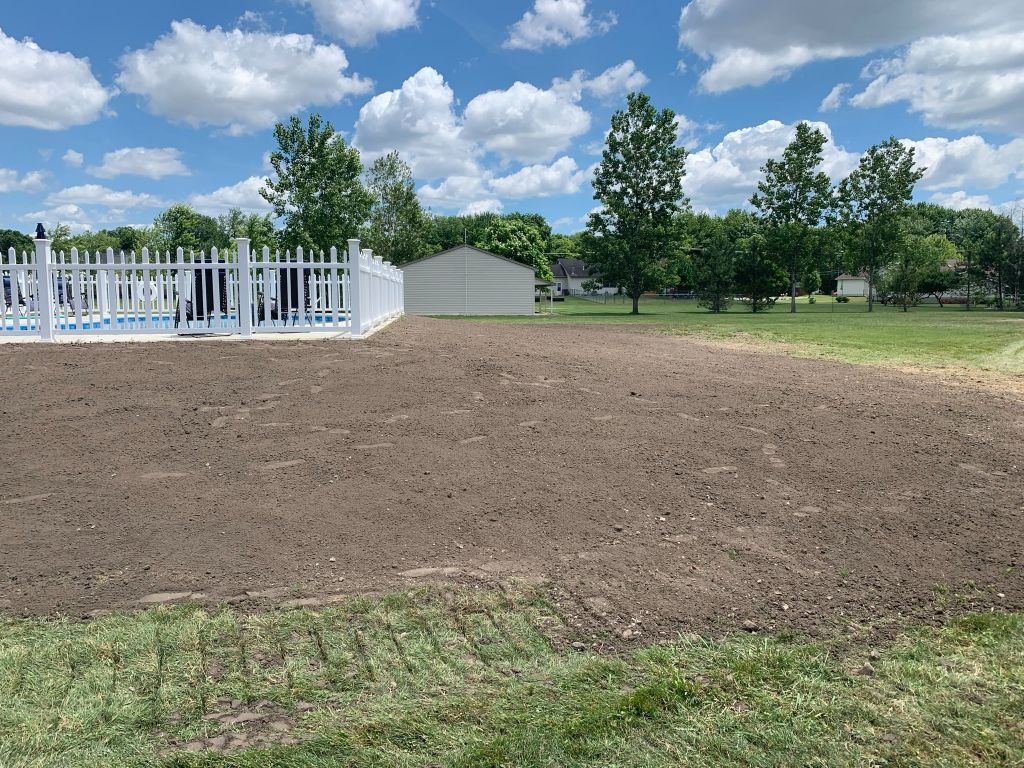 Freshly tilled dirt field beside a white fence, trees, and a small shed under a cloudy blue sky