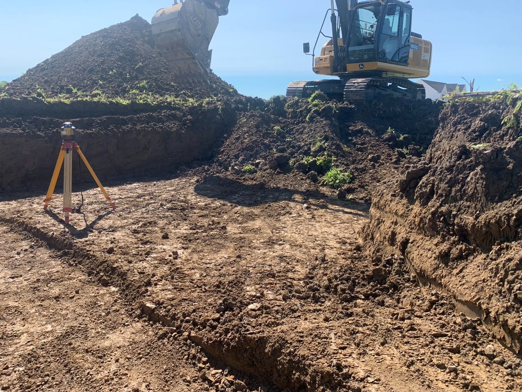 Excavator on a muddy construction site with dirt mounds and a survey tripod under a clear sky