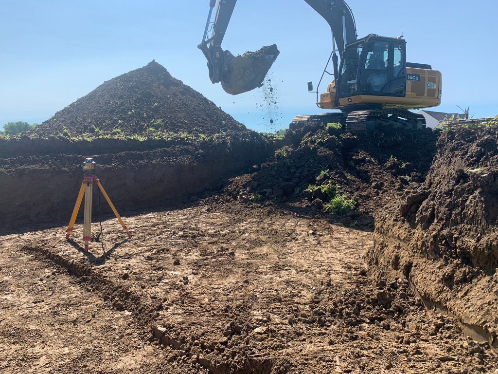 Excavator dumping dirt at a construction site with a large soil pile and a tripod survey marker