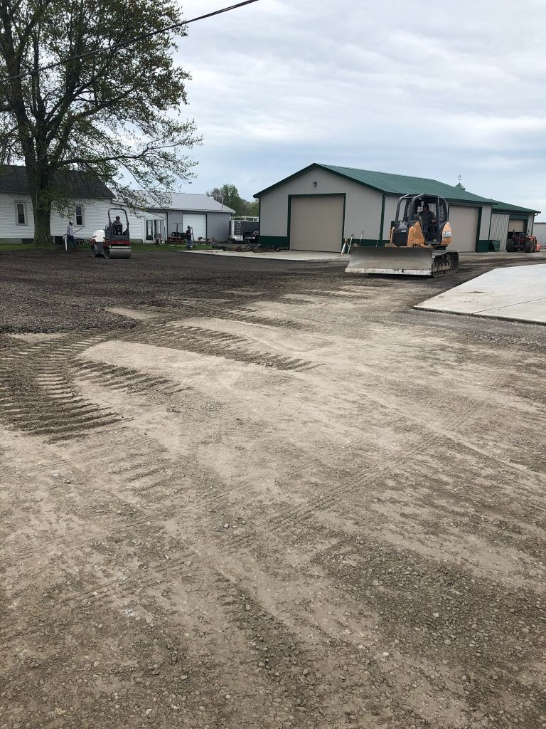 Gravel lot with a house and garage, construction equipment, and a tree on an overcast day