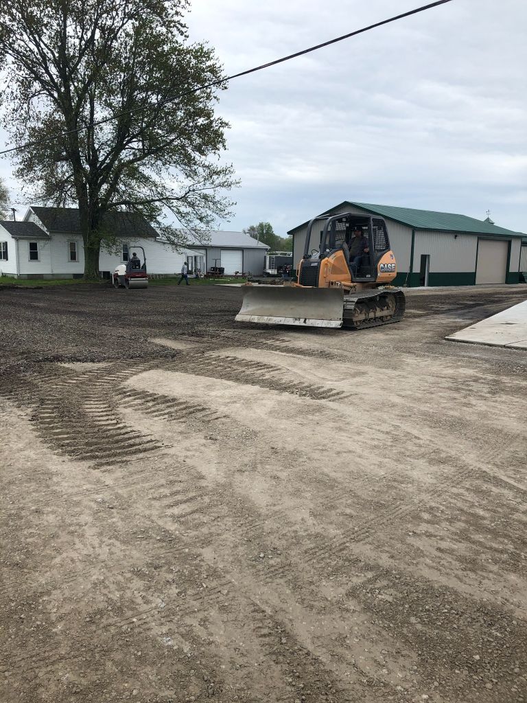 Construction equipment leveling a dirt lot near houses and a large building under an overcast sky