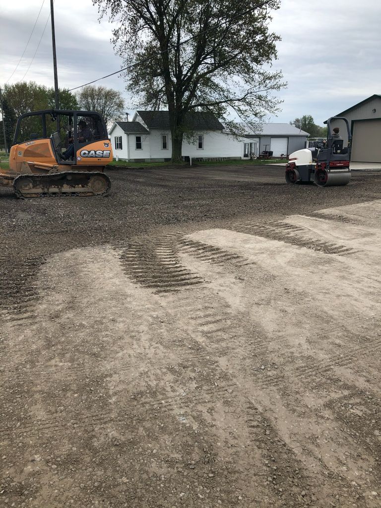 Two excavators grading a dirt lot in front of a white house and tree under an overcast sky