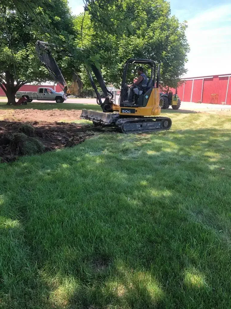 Mini excavator mowing grass under a tree in a park-like yard.