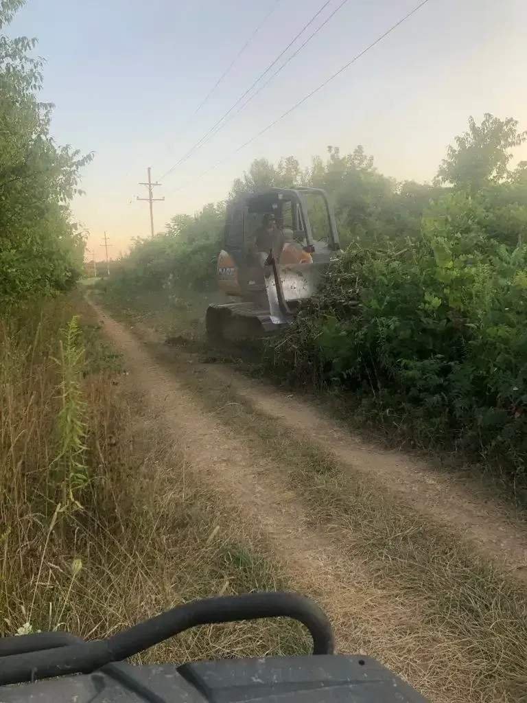 Tracked excavator clearing bushes beside a dirt path in a hazy rural field