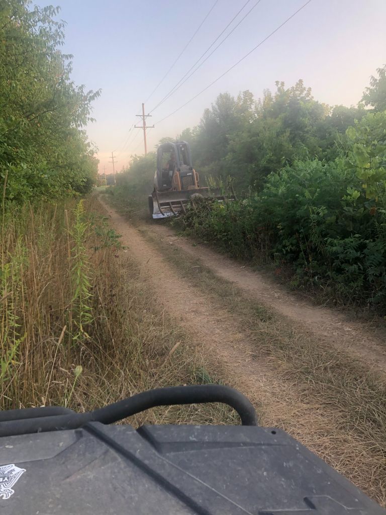 Dusty dirt road lined with trees, viewed from an off-road vehicle behind a tractor ahead at dusk