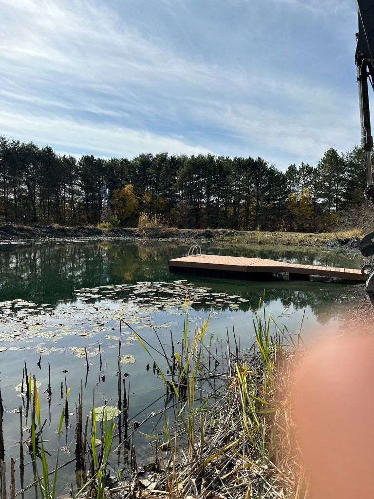 Calm lake with a small dock, reeds in the foreground, and trees lining the far shore under a cloudy sky