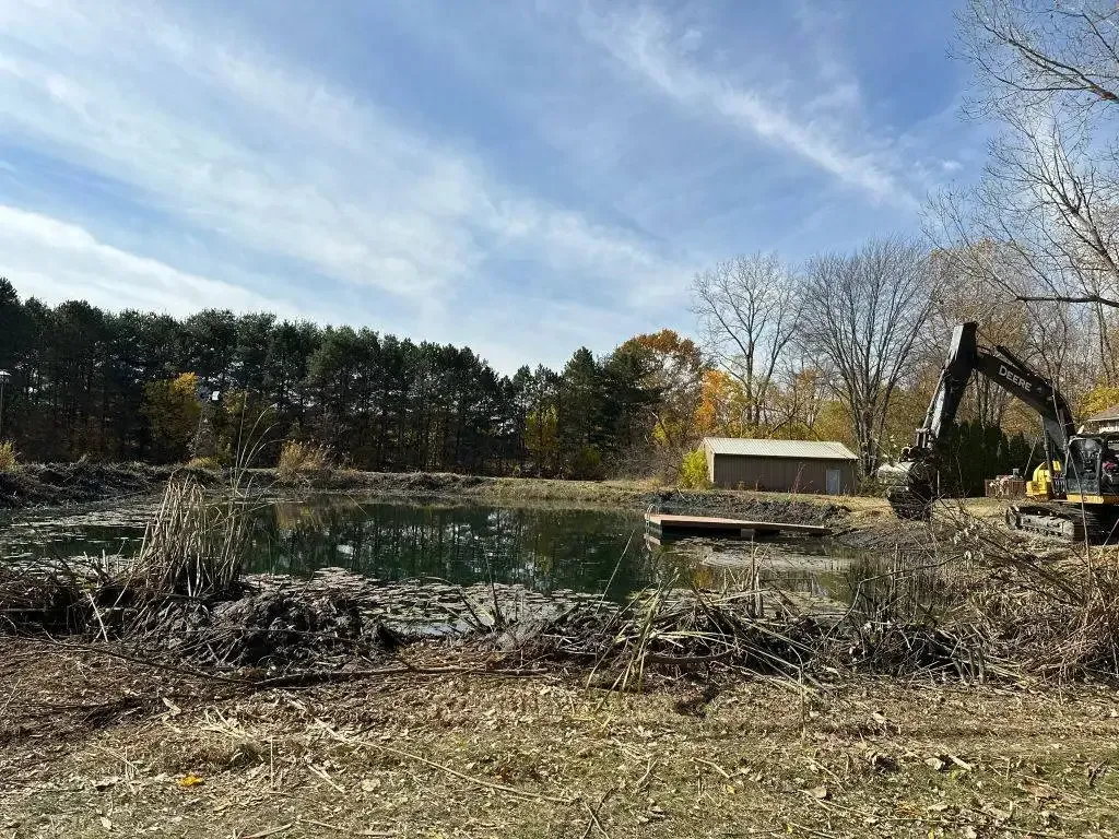 Excavator clearing debris at a site beside a pond under a blue sky, with trees in the background