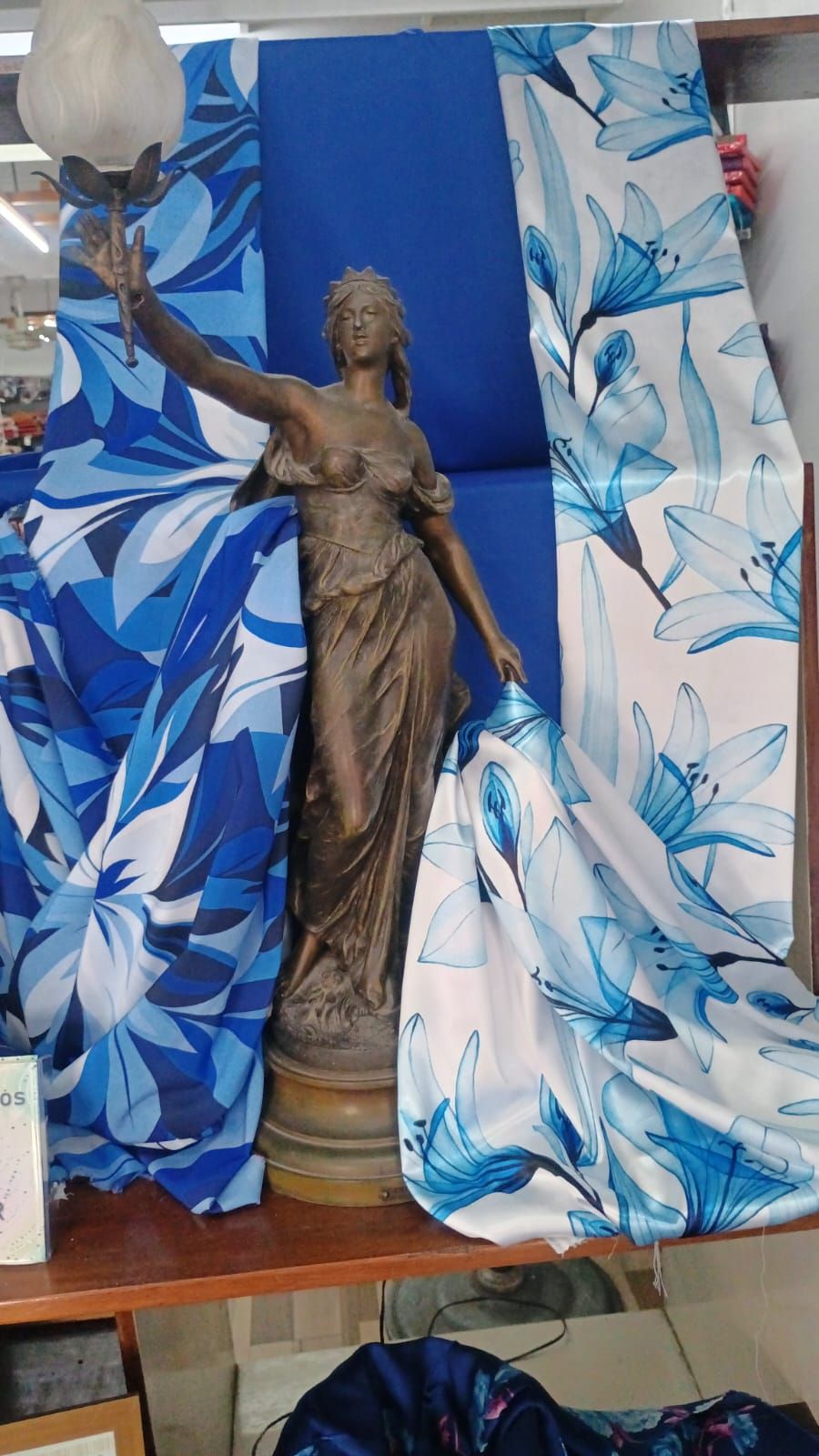 Una estatua clásica en tonos bronce que sostiene una pequeña lámpara, enmarcada por una tela floral azul y blanca