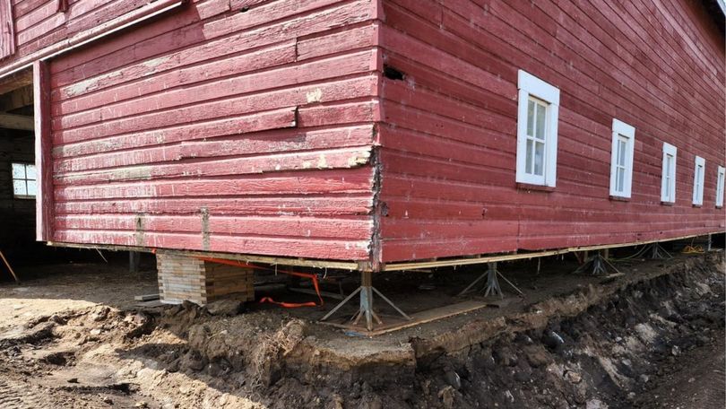 A red wooden barn elevated on metal jack posts and timber cribbing above an excavated dirt foundation.