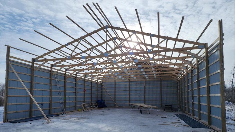 A large pole barn under construction with a wooden roof truss frame and metal siding panels on a snowy day.