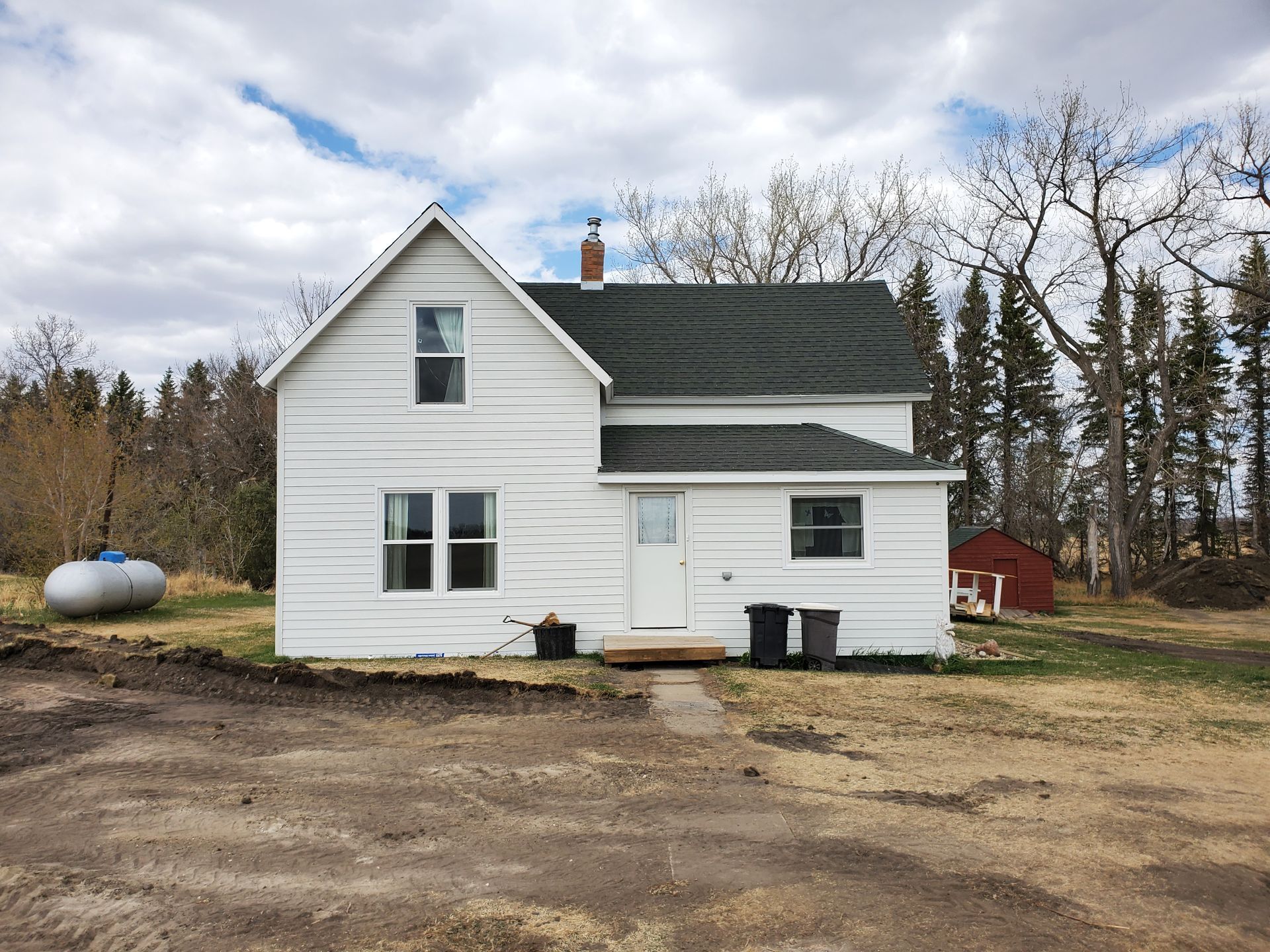 White farmhouse with green roof on a dirt lot, backed by trees under a cloudy sky