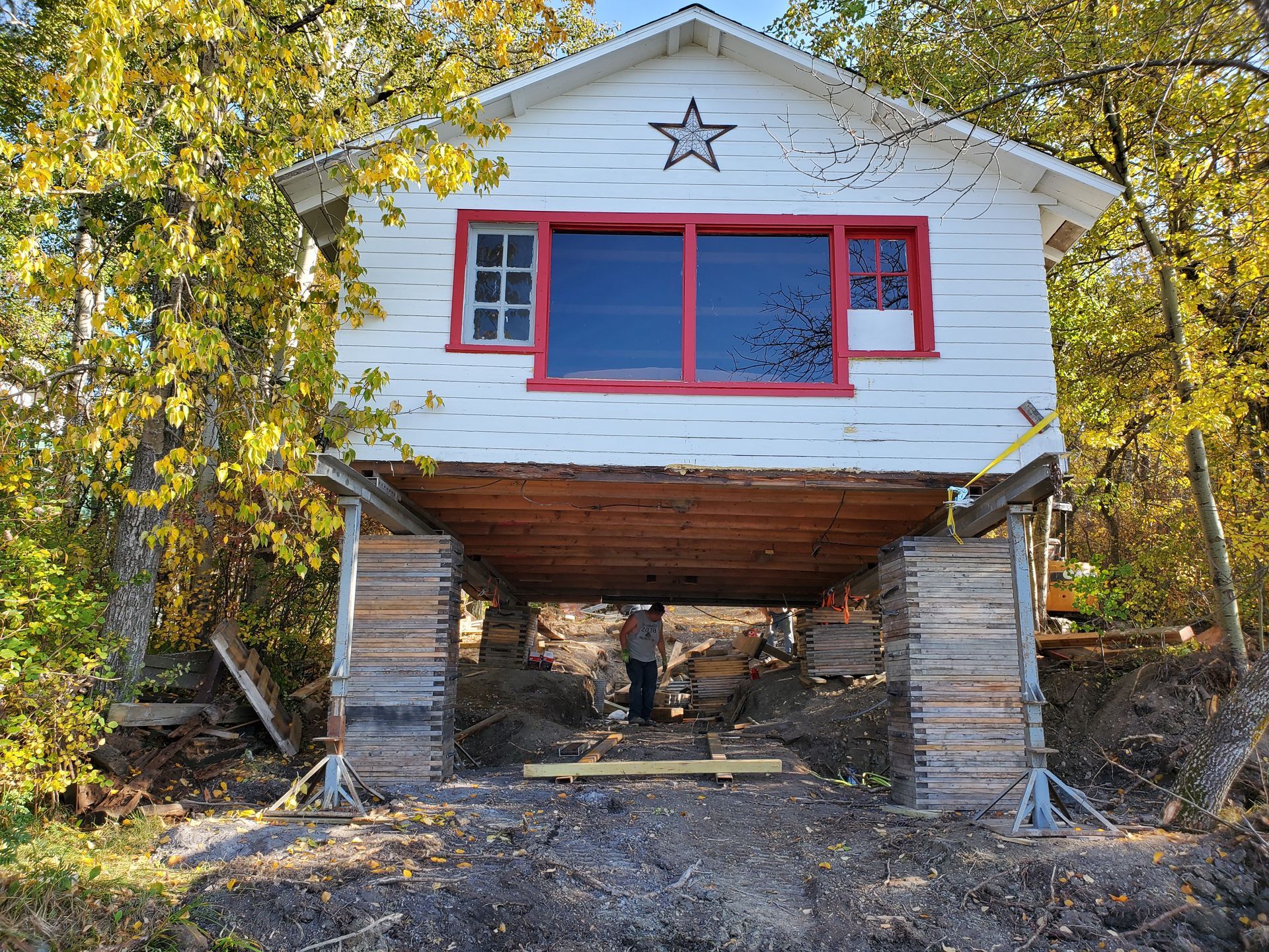 White cabin with red-trimmed windows and star, raised on stone supports in a wooded hillside.