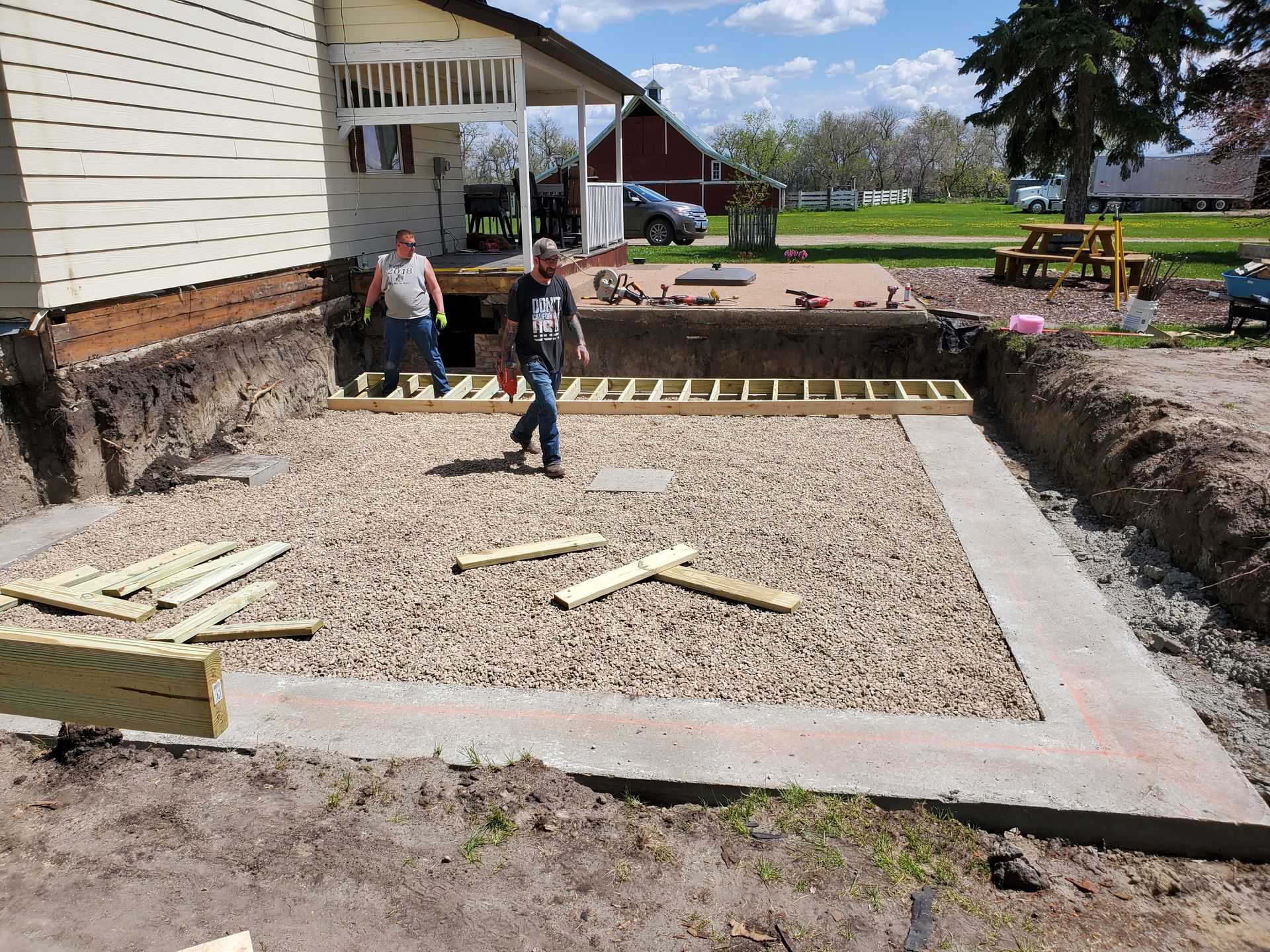 Construction site with two workers pouring and leveling a gravel foundation beside a house
