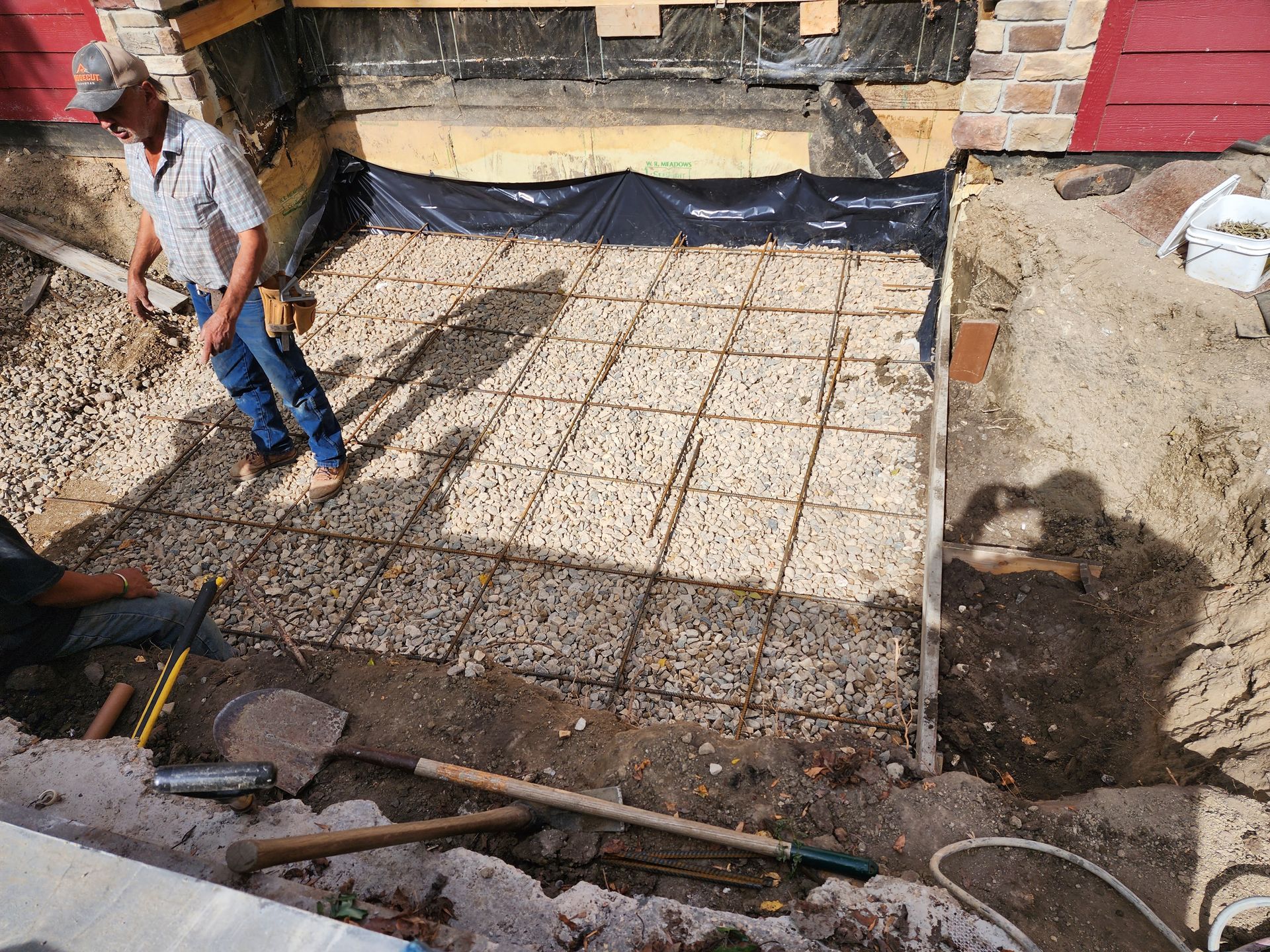 Construction site with excavated foundation, rebar grid, black waterproofing, and worker standing at left.