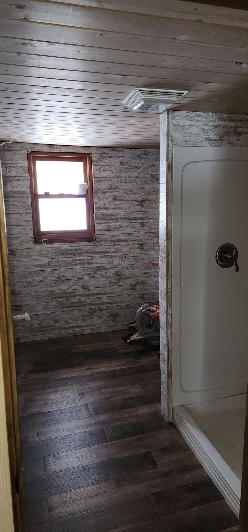 Narrow hallway with wood ceiling, window, brick wall, and white shower stall on the right.