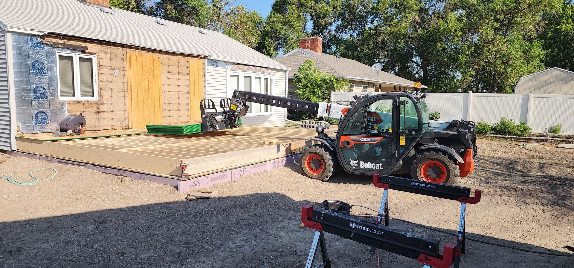 Small backhoe loader on gravel near a house with boarded windows and a fenced yard