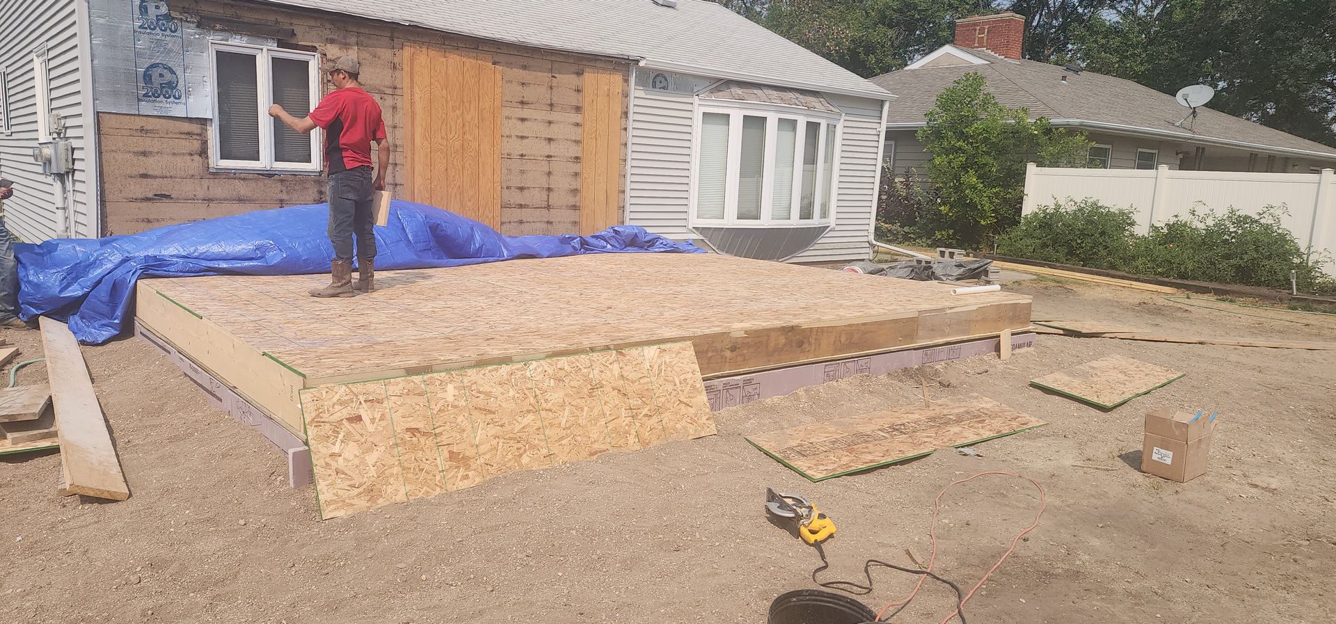 Worker on a roof under construction beside wooden framing and blue tarp near a house.