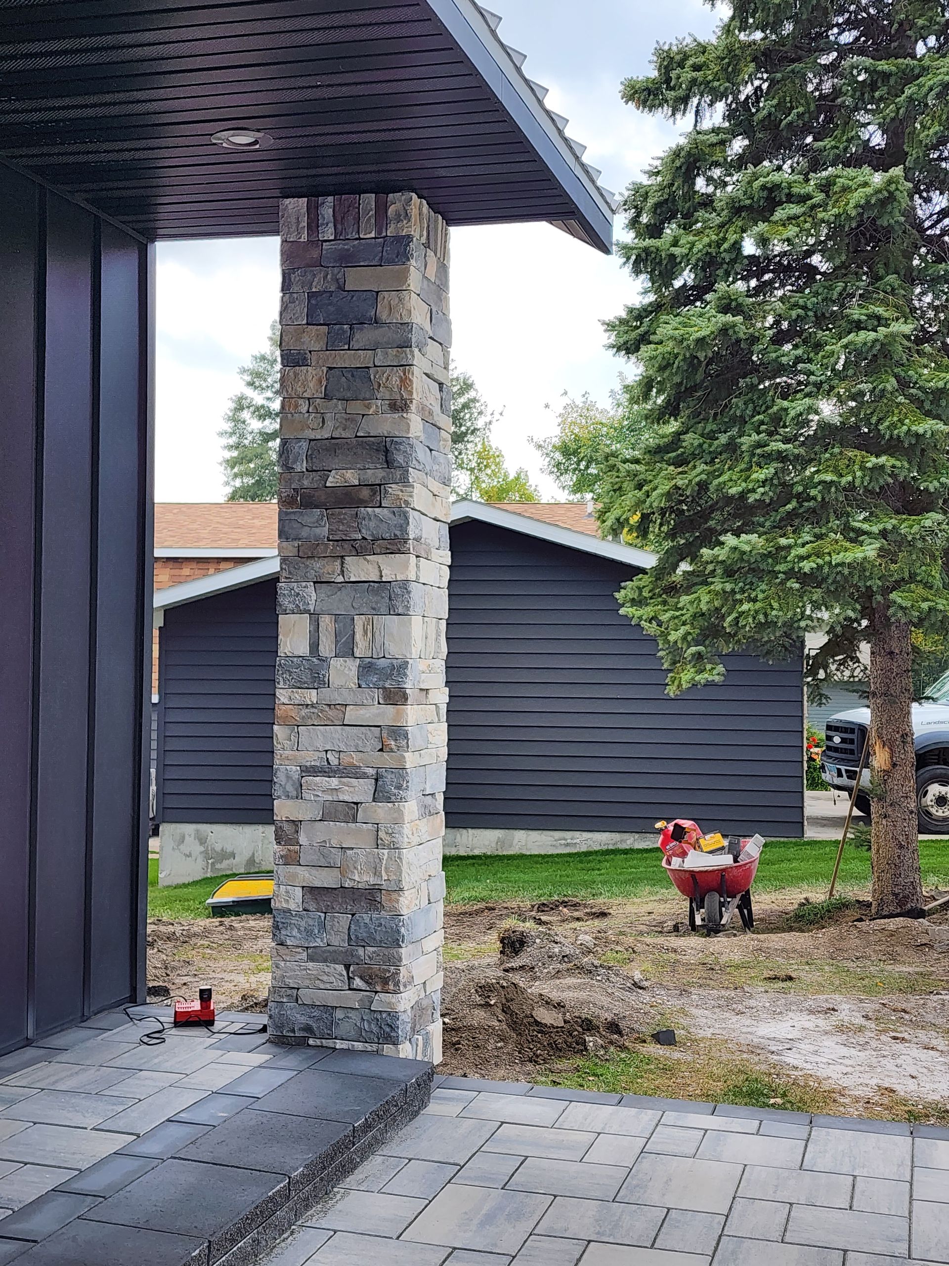 Stone porch column beside dark siding, with a red wagon near a tree in a gravel yard.