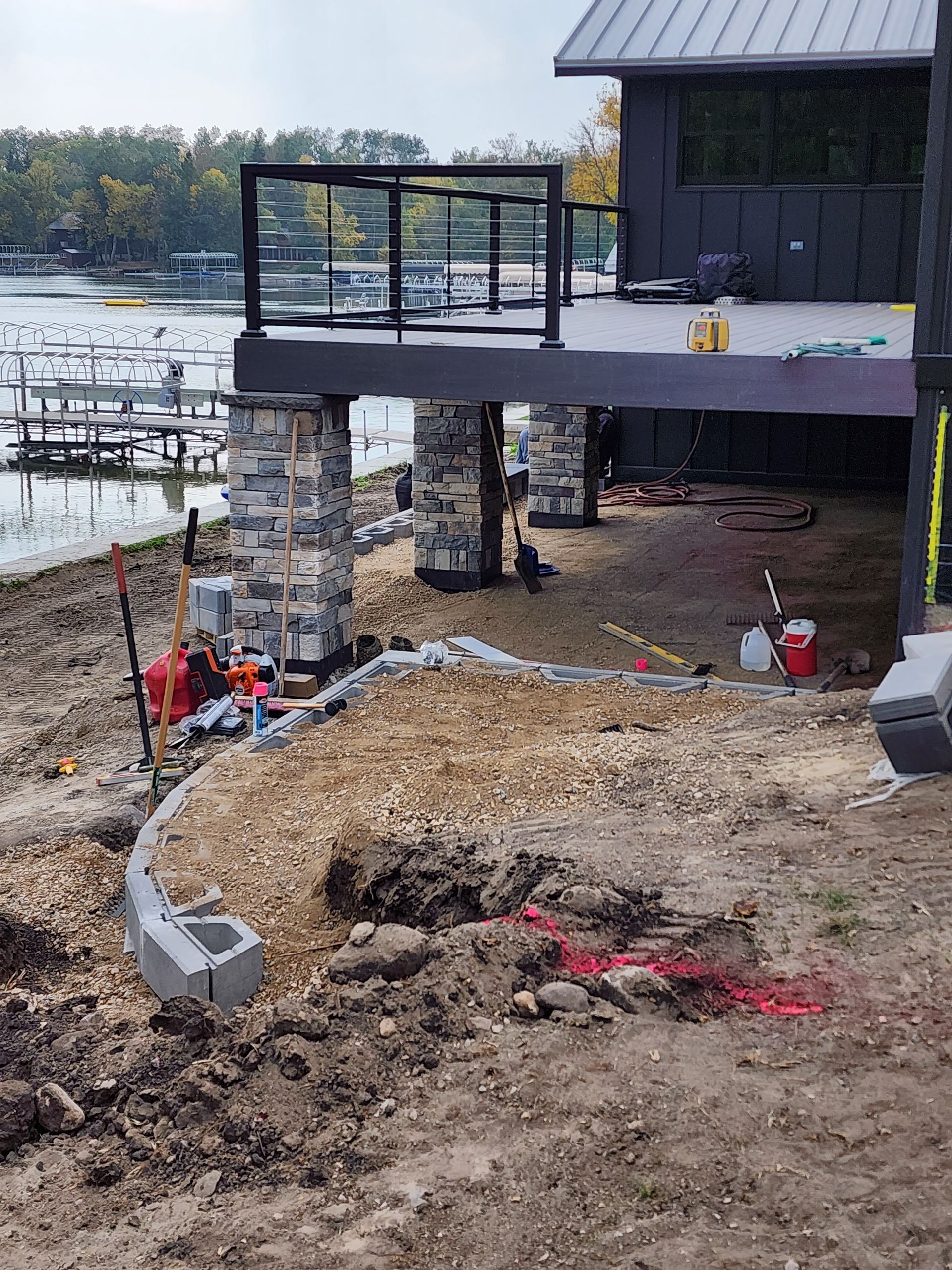 Riverside deck under construction with stone footings, excavated ground, and tools beside the water