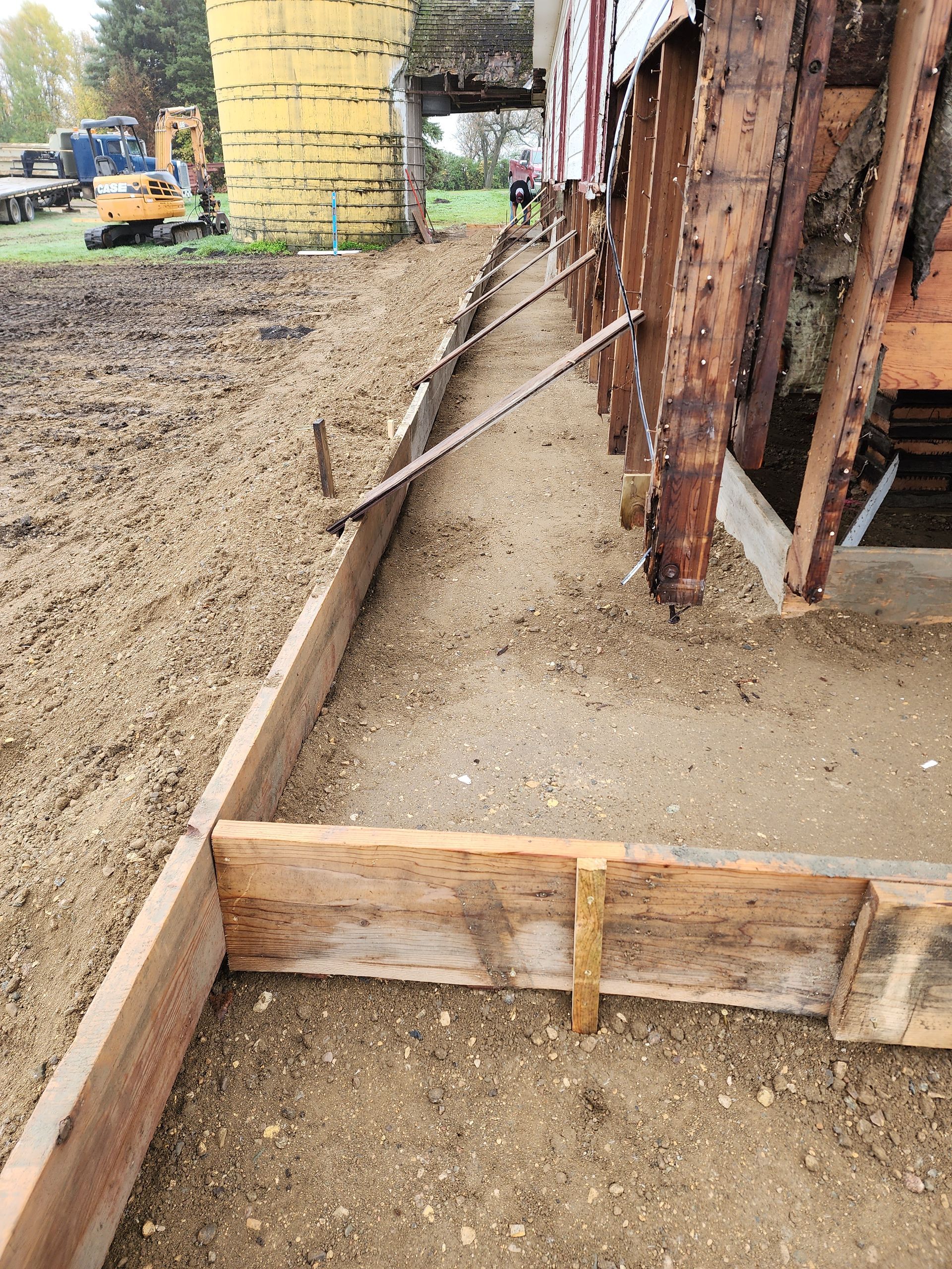 Construction site with dirt foundation, wooden forms, and steel support beams beside a building under construction
