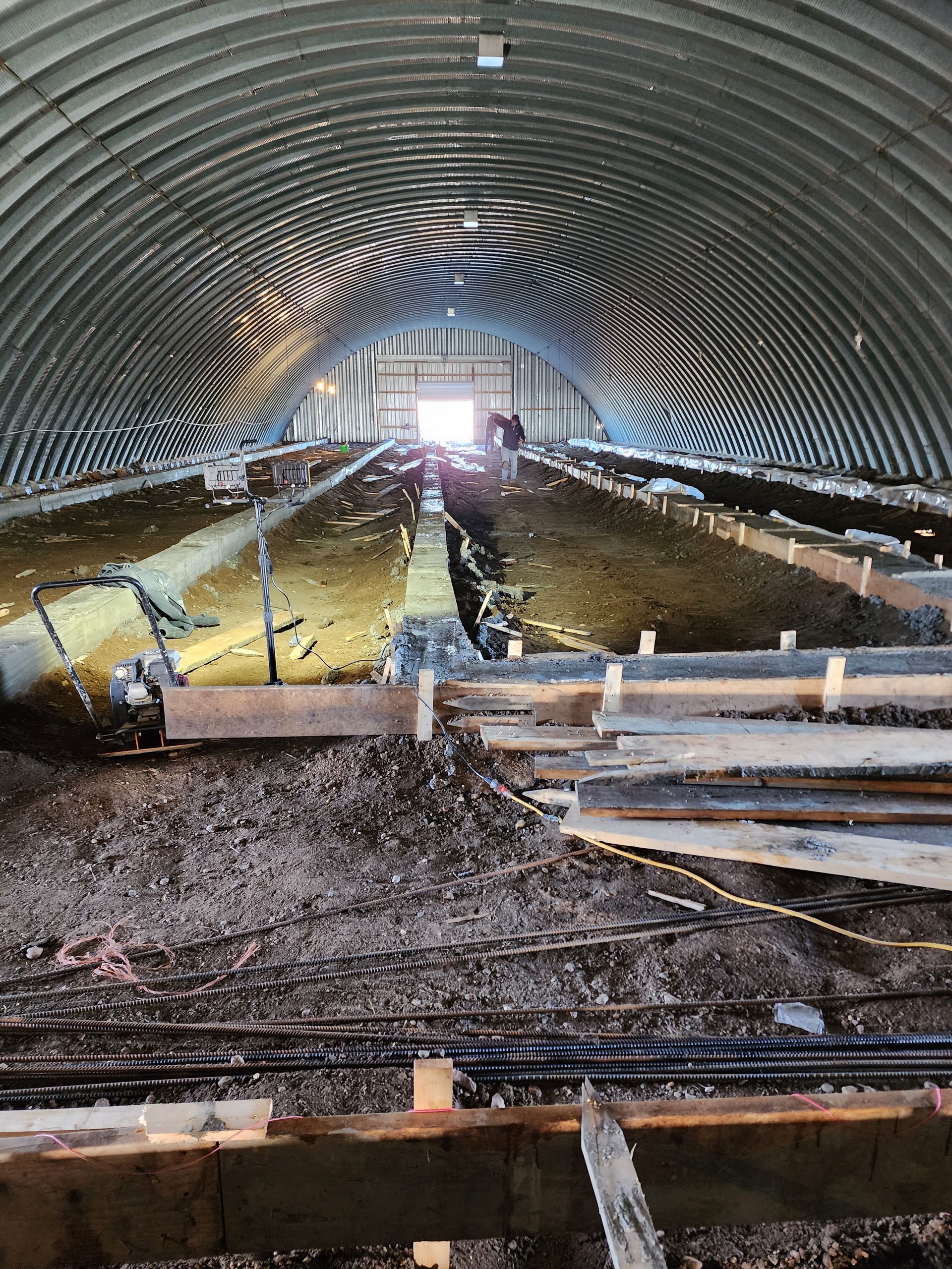 Inside a corrugated tunnel under construction, with wet ground, barriers, and daylight at the far end.