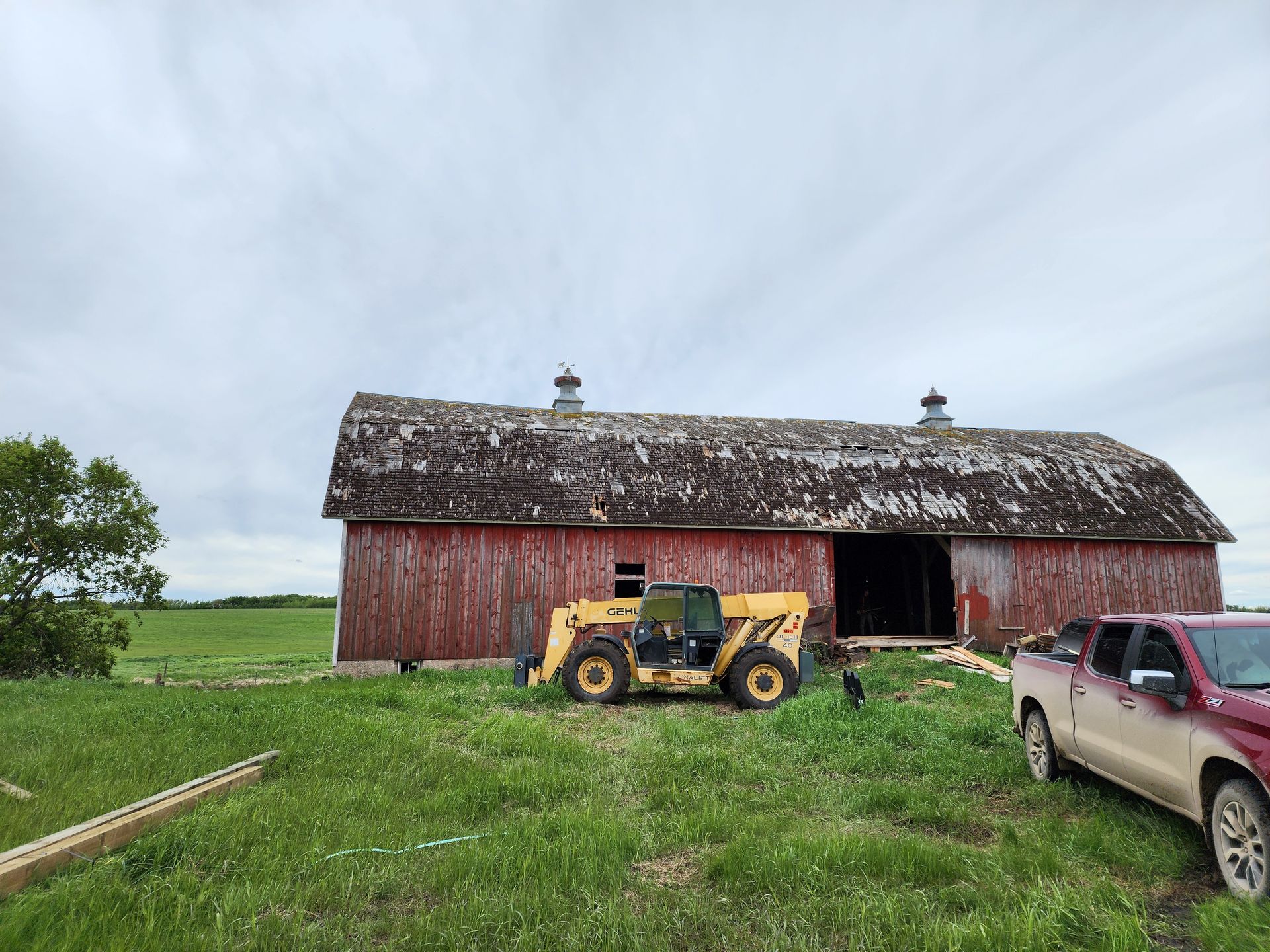 Yellow tractor beside a weathered red barn with a pickup truck in a grassy field under a cloudy sky