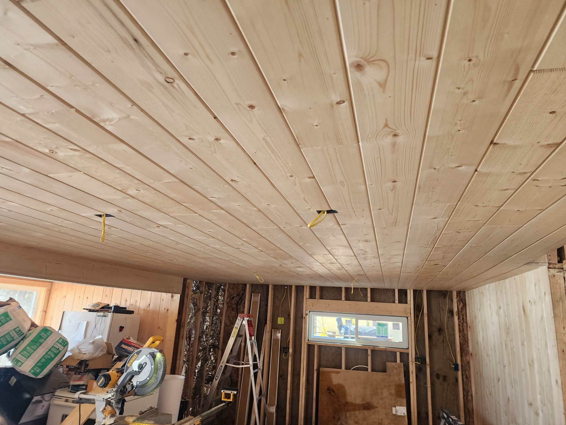 Partially finished room with wood-paneled ceiling, exposed walls, ladders, and construction tools