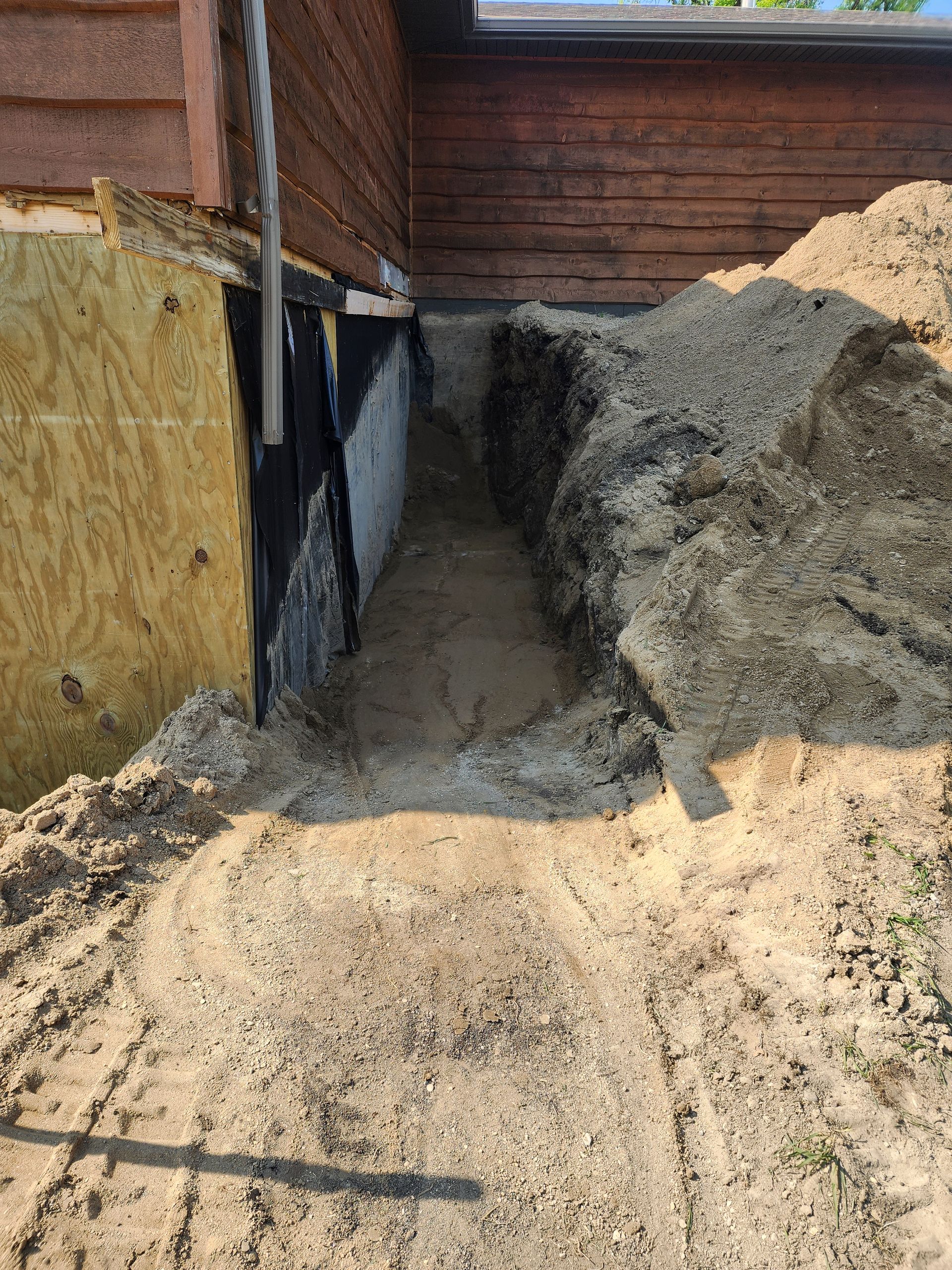 Narrow dirt trench beside a brick building wall, with soil piles on both sides and exposed foundation.