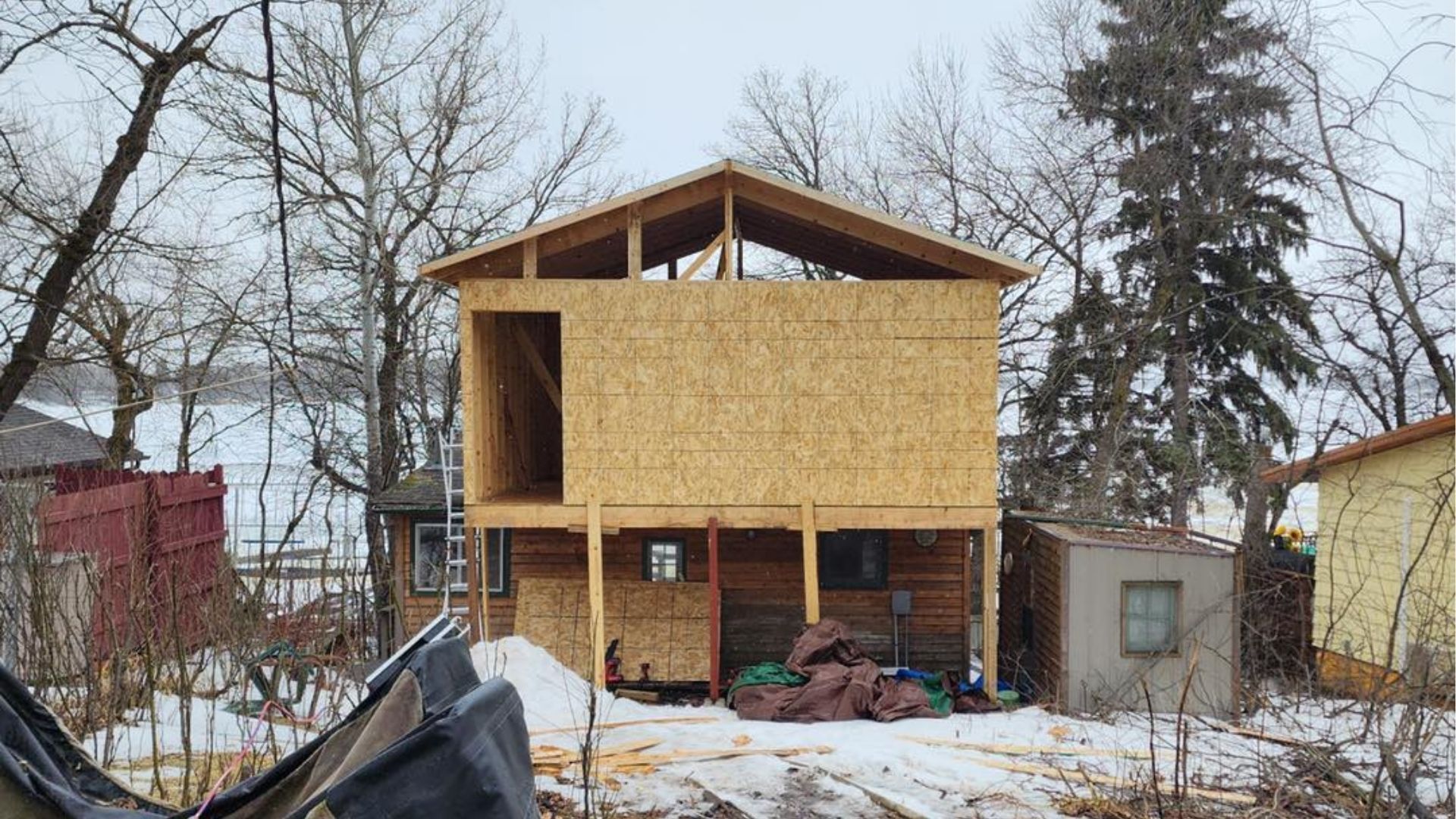 A two-story building under construction with OSB-sheathed walls and an open gable roof set among trees in winter.