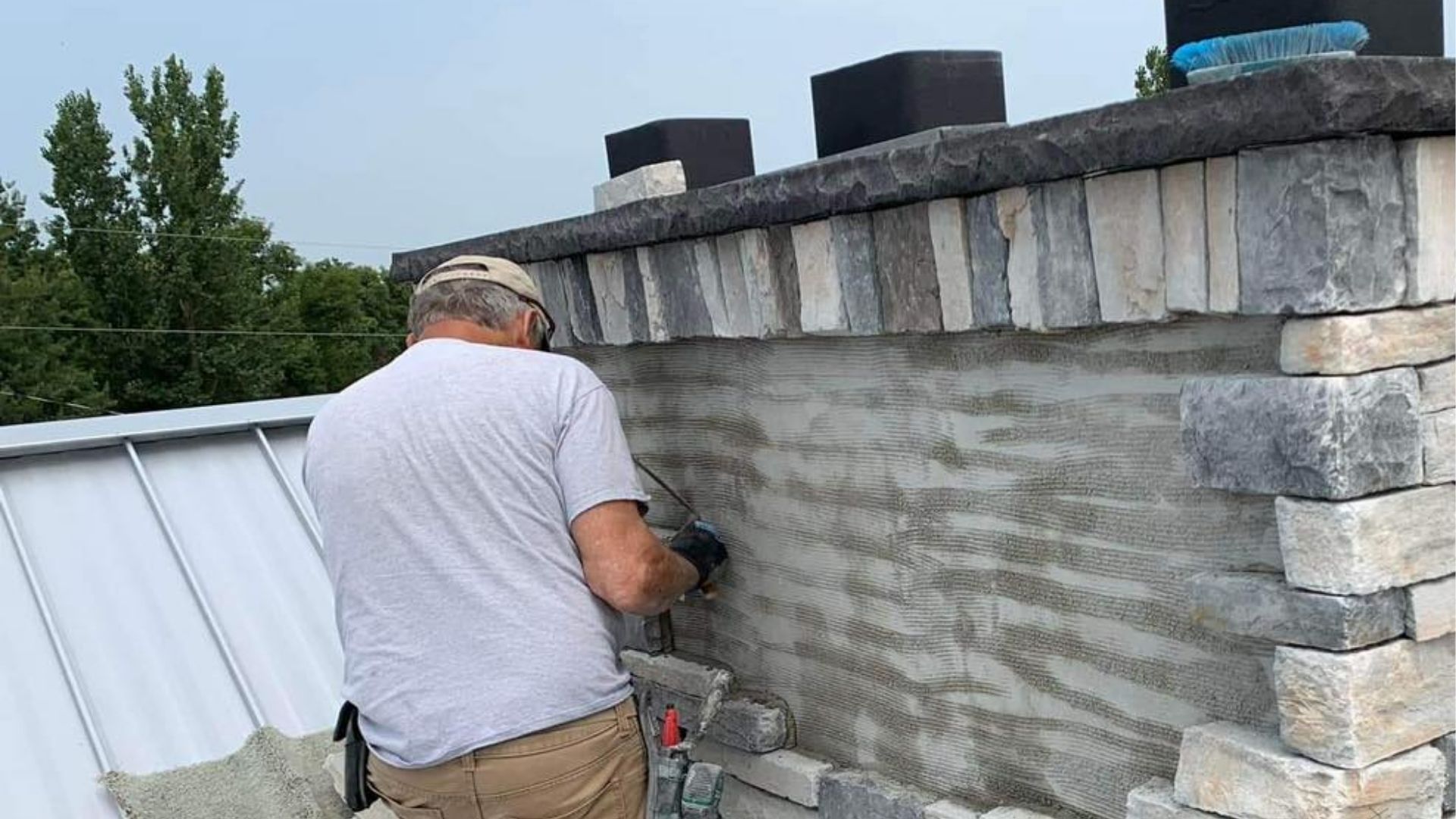 A worker applies stone veneer to the side of a chimney on a metal roof.